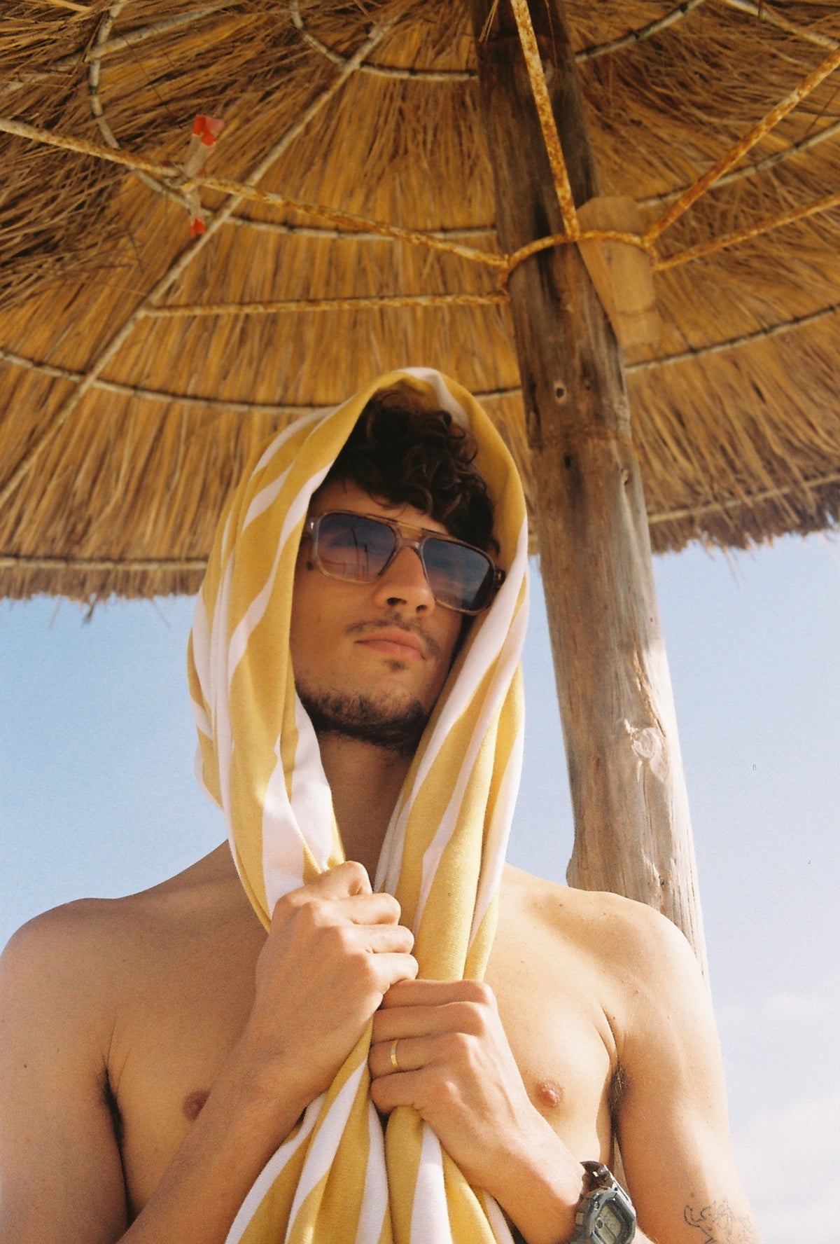 A shirtless man in sunglasses with a yellow striped towel over his head, under a straw beach umbrella