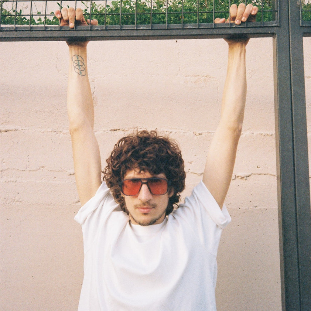 A man with curly brown hair and steampunk reddish-tinted sunglasses wears a white t-shirt and holds a black metal grid fence overhead against a light beige wall.