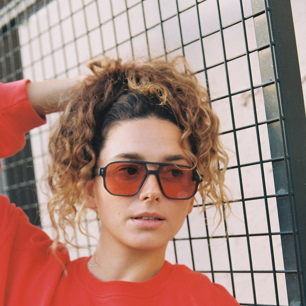 A woman with curly light brown hair wearing oversized square sunglasses with red-tinted lenses and a bright red sweatshirt, posing in front of a black metal grid fence in the Balearics 