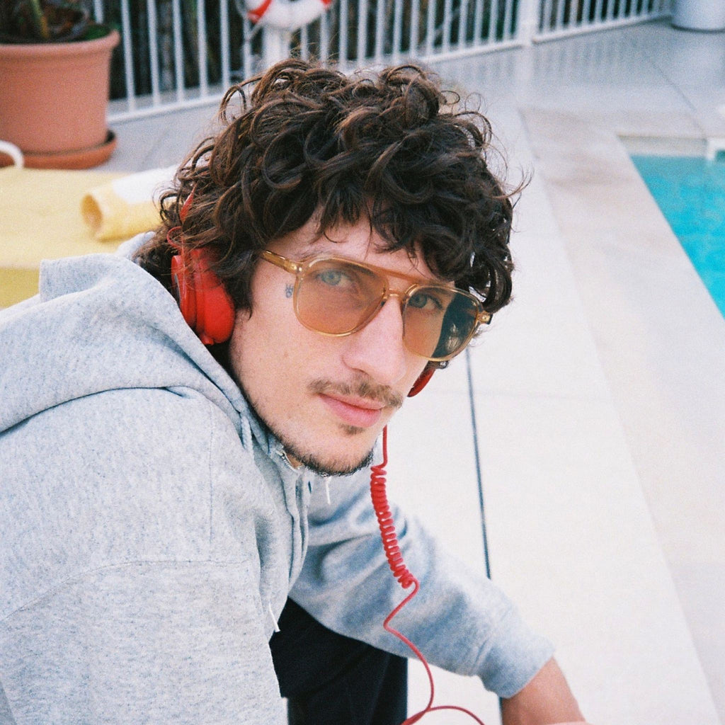 A man with curly dark hair wearing a light gray hoodie, gold-framed aviator sunglasses, and red over-ear headphones with a red coiled cord, leaning by a swimming pool.
