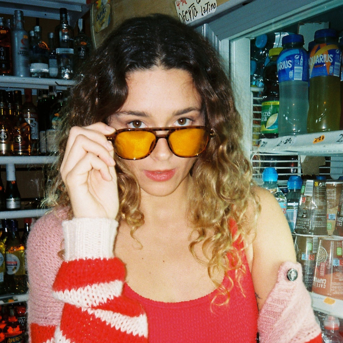 A woman with curly hair wearing yellow tortoise -tinted sunglasses, a red tank top, and a red-and-white striped knit cardigan, standing in front of a refrigerator stocked with bottled drinks in what looks like a convenience store in Ibiza island.
