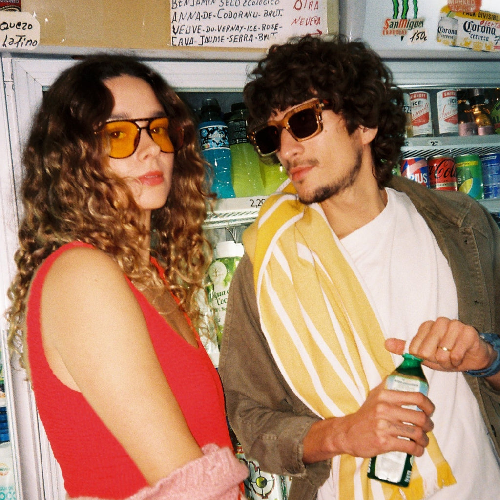 A man and a woman posing in front of a refrigerator stocked with drinks in Ibiza. The woman wears orange-tinted sunglasses, a red top, and a pink and red striped knit sweater draped over her arm.