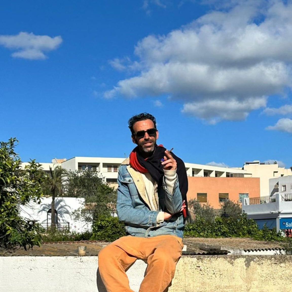 A man wearing sunglasses, a blue denim jacket, a cream and red scarf, and orange pants sits casually on a low concrete wall outdoors under a bright blue sky with clouds In Ibiza