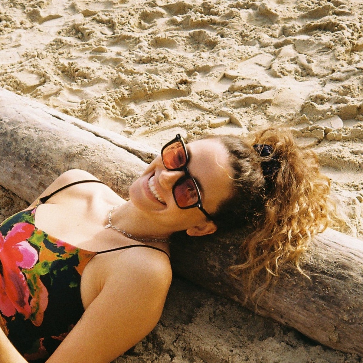 A woman wearing a colorful floral swimsuit and sunglasses relaxes on a sandy beach next to a large log.