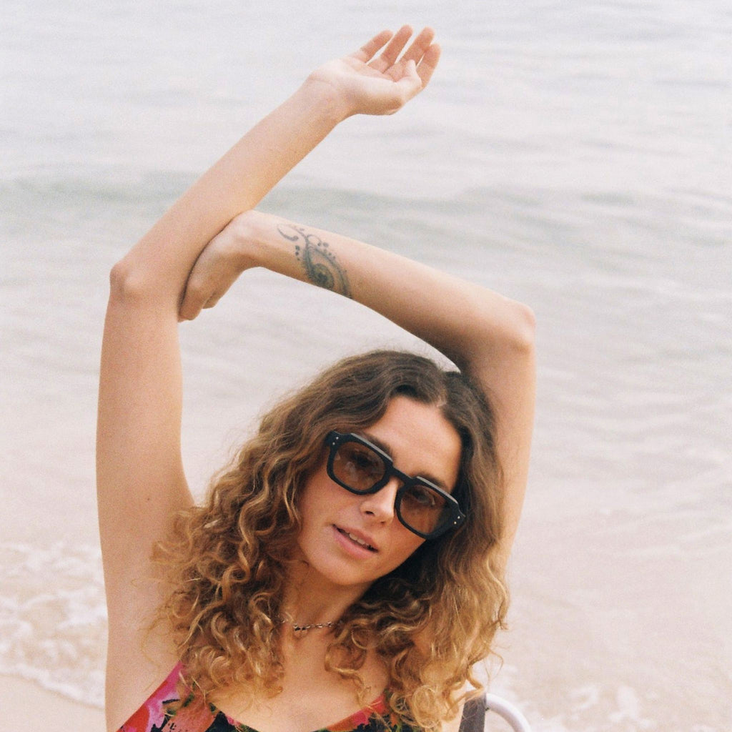 A woman with curly hair and a tattoo on her arm wearing dark sunglasses and a colorful dress, posing with her arms raised above her head on a beach in ibiza with the ocean in the background.