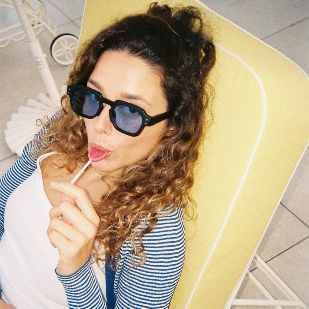 A woman with curly brown hair wearing black sunglasses, a blue and white striped cardigan over a white top, and a lollipop, sitting in a yellow lounge chair.