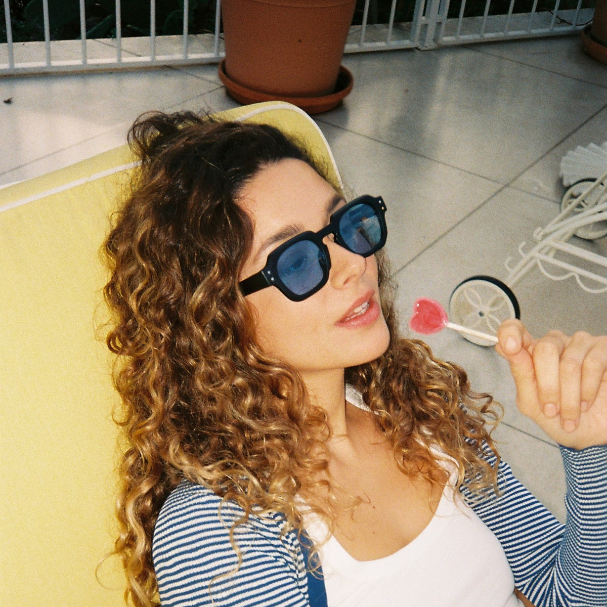 A woman with long curly brown hair wearing black sunglasses, a blue and white striped cardigan over a white tank top, and a yellow hat, sitting on a yellow lounge chair in an outdoor patio setting.