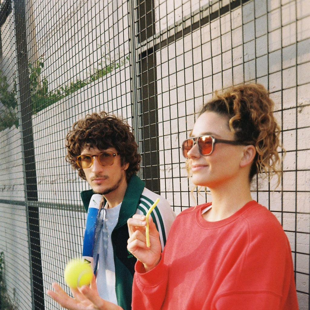 Two people on a tennis court wearing athletic apparel. The person in the foreground wears a bright red sweatshirt and round sunglasses while holding a tennis racket.
