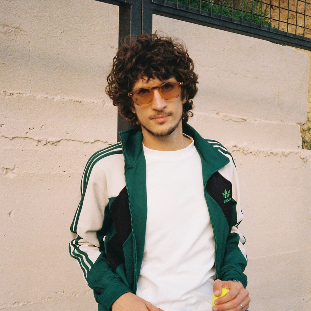A man wearing a green and white Adidas track jacket, white t-shirt, and sunglasses sits against a light-colored brick wall holding a blue pickleball paddle.