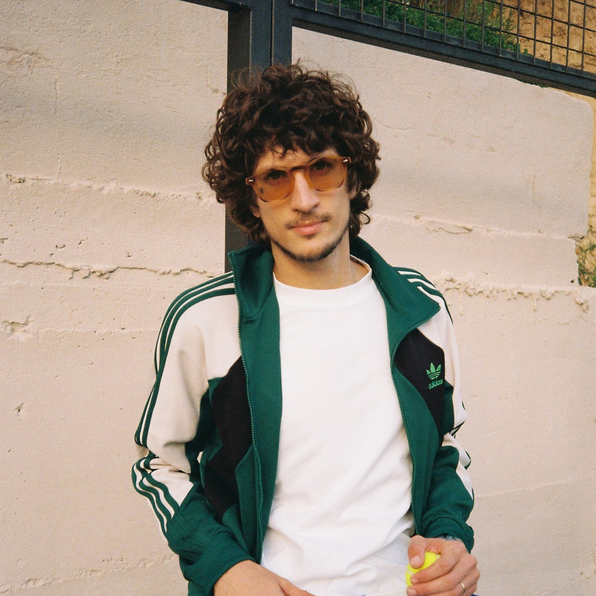 A man wearing a green and white Adidas track jacket, white t-shirt, and sunglasses sits against a light-colored brick wall holding a blue pickleball paddle.