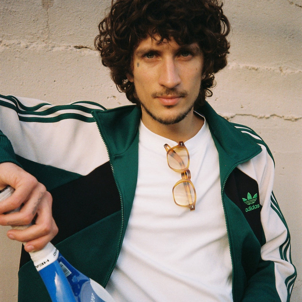 A man with curly dark hair wearing a green and white Adidas track jacket, white t-shirt, and holding a blue and white tennis racket and a yellow tennis ball, posing against a textured concrete wall.