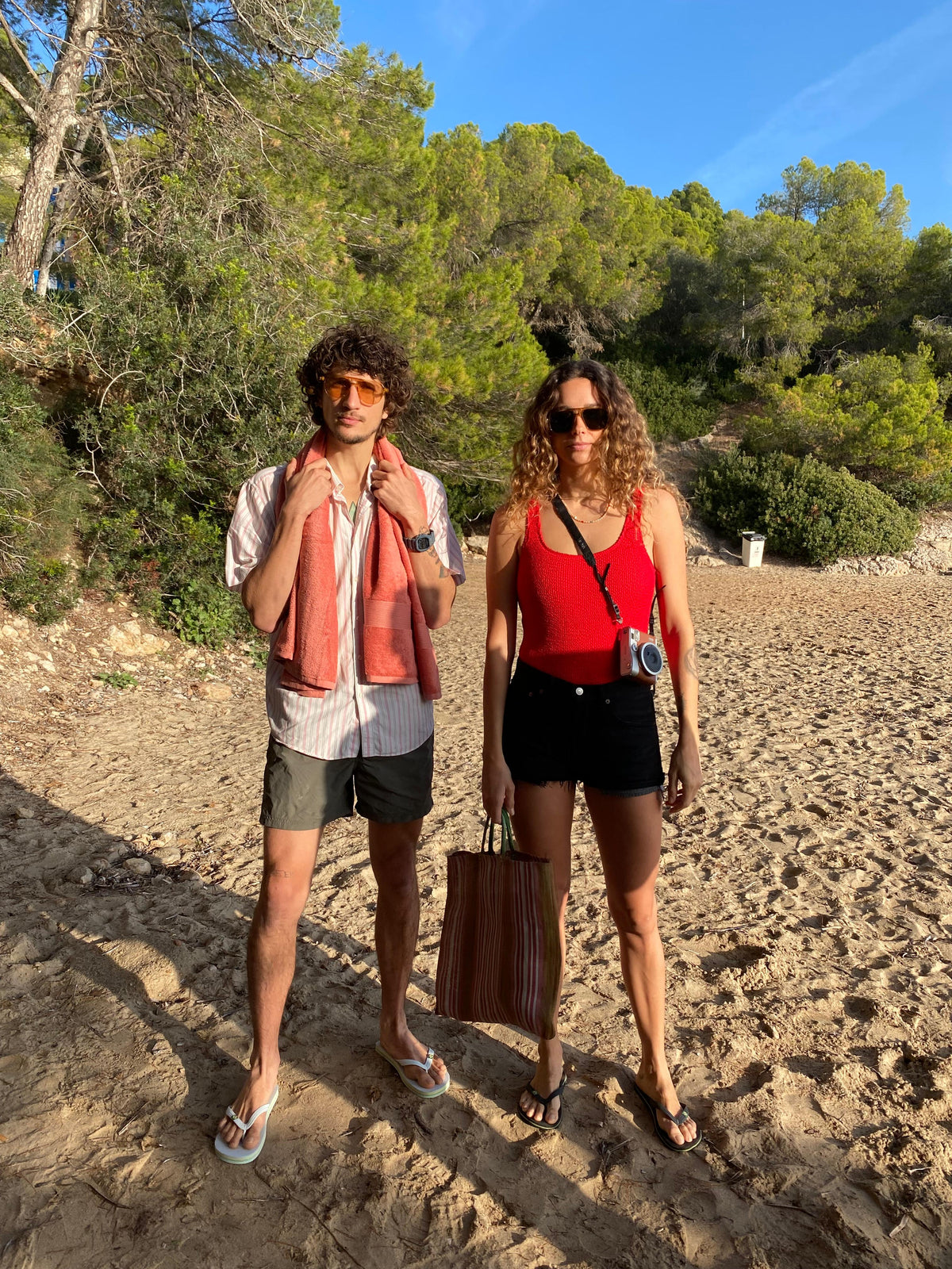 A man and a woman in sunglasses standing on a sandy beach under pine trees in the sun