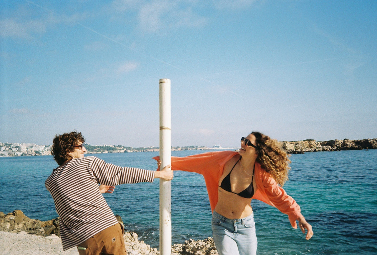 Two friends in sunglasses by a sailing mast with the sea behind them