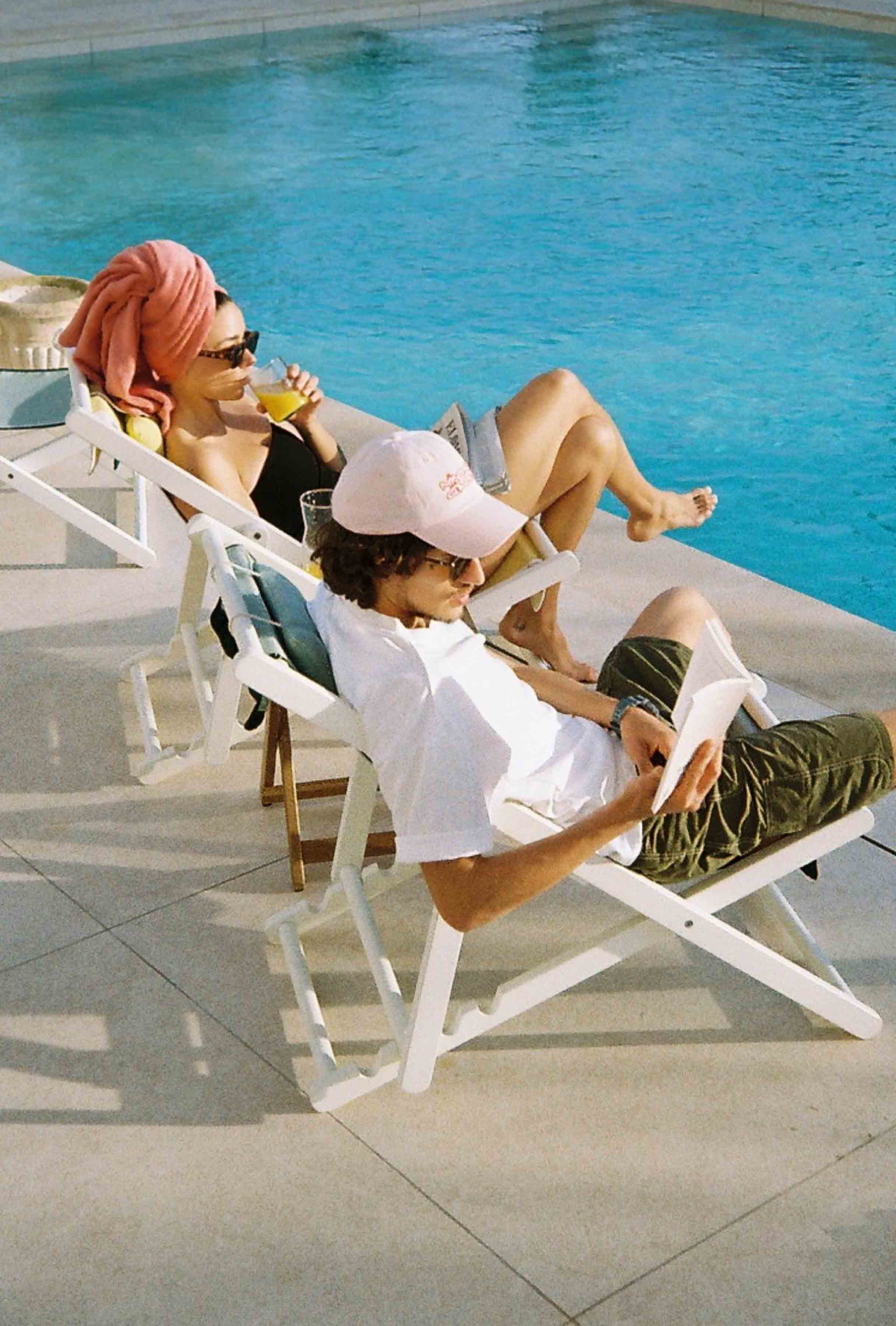 Two people with sunglasses relaxing by a poolside on lounge chairs.